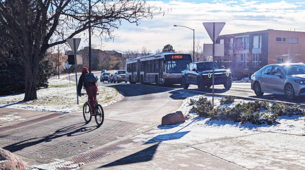 The image shows a bicyclist riding across a turn lane off Broadway, while a line of cars, trucks and buses travels on Broadway.