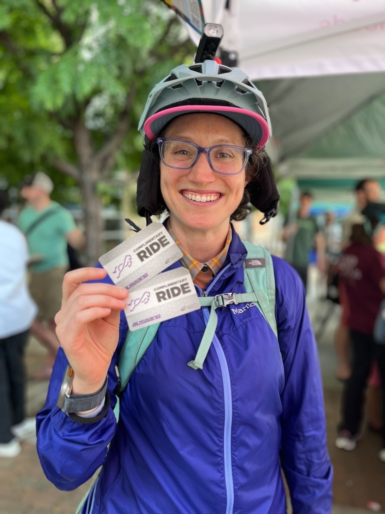 A person wearing glasses and a bicycle helmet holds up two bus tickets and smiles
