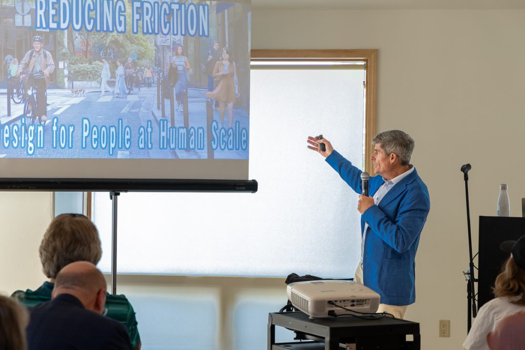 A man in a blue sport coat gestures to a screen showing a slide that reads, "Reducing Friction for People at Human Scale."