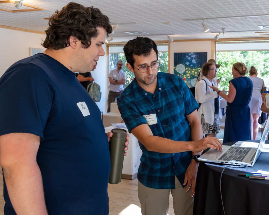 Two light-skinned men with dark hair stand together looking at a laptop computer, while one shows the other something on the screen.