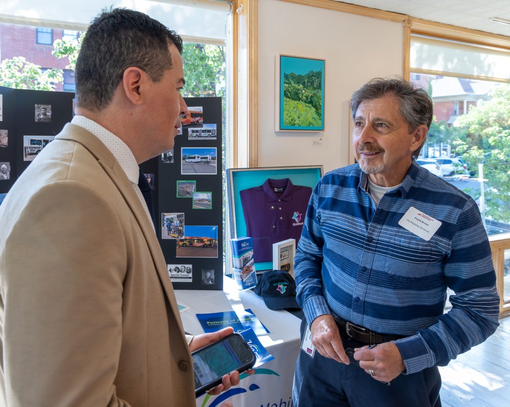 Two men face each other in conversation, in front of a table displaying photographs and materials.