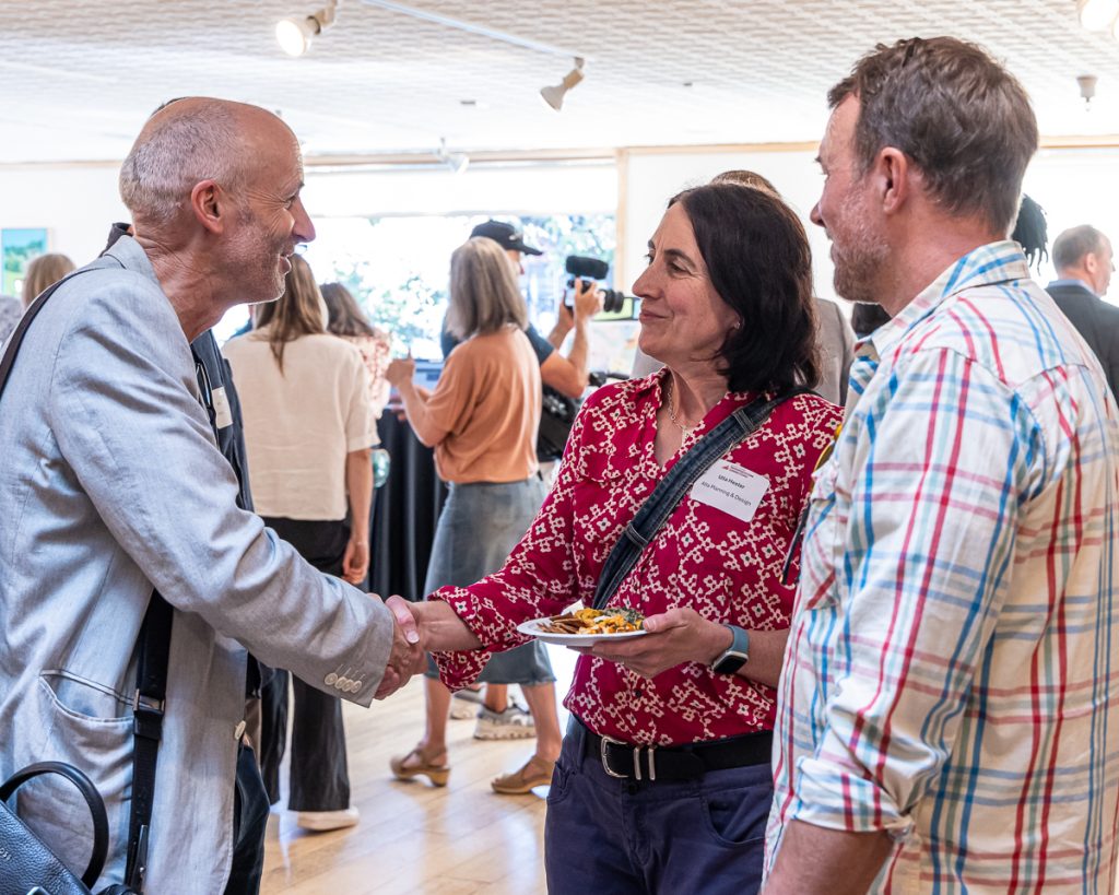 A man and a woman shake hands and smile at each other in a brightly lit room full of people.