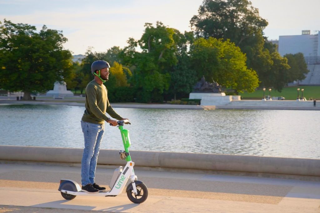 A person rides a Lime e-scooter on a concrete path next to a manmade reservoir.