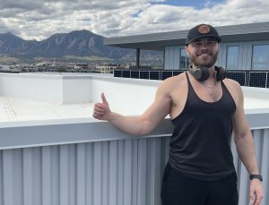 A man wearing a baseball cap, a black tank top and headphones smiles and gives a thumbs-up to the photographer at his apartment building.