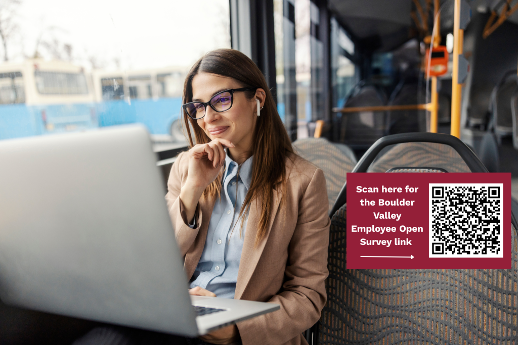 A woman in business clothing sits on a bus looking at her laptop computer and smiling.