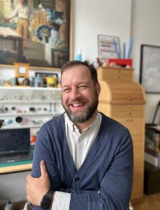A portrait of Jon Jon Wesolowski - a light-skinned, bearded man wearing a white shirt with a blue cardigan sweater, smiling.