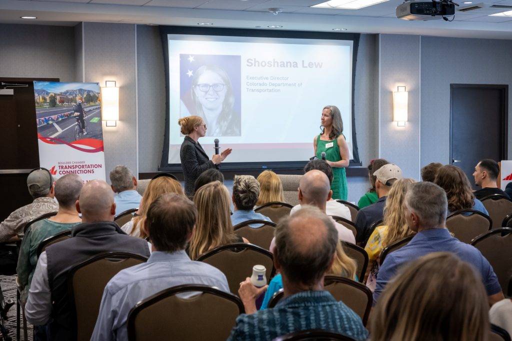 An image of a conference room with two people standing in front of a screen that shows a picture of Shoshana Lew